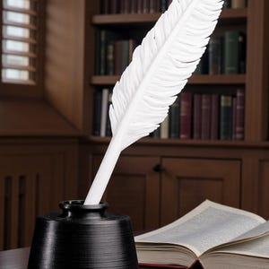 May include: A white feather quill pen in a black inkwell, set on a wooden desk. The quill is prominently displayed, with a detailed feather design. An open book and a bookshelf filled with books are in the background.