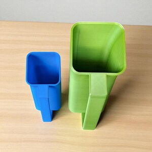 May include: Two plastic scoops, one blue and one green, sit side-by-side on a light wood surface. The scoops have a square top and a handle. The green scoop is larger than the blue scoop.
