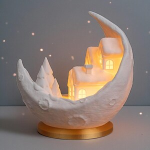 May include: A white crescent moon-shaped decorative lamp with a miniature snow-covered village inside. The village features illuminated houses and trees. The lamp sits on a gold-colored base, creating a warm, inviting ambiance. The overall theme is winter and holiday decor.