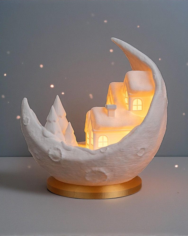 May include: A white crescent moon-shaped decorative lamp with a miniature snow-covered village inside. The village features illuminated houses and trees. The lamp sits on a gold-colored base, creating a warm, inviting ambiance. The overall theme is winter and holiday decor.