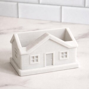 May include: A white, house-shaped container with a pitched roof, two square windows, and a front door. The container is made of a solid, opaque material and sits on a rectangular base. The windows and door are recessed.