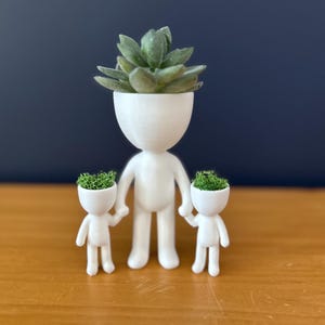 May include: A white, 3D-printed planter shaped like a person, holding the hands of two smaller planters. Each planter has a head filled with green plants. The background is a dark blue wall and a wooden surface.