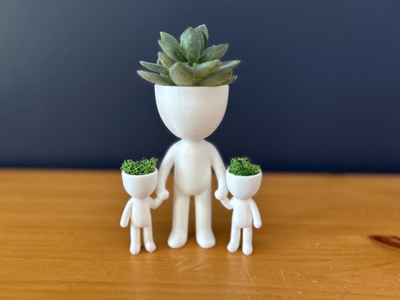 May include: A white, 3D-printed planter shaped like a person, holding the hands of two smaller planters. Each planter has a head filled with green plants. The background is a dark blue wall and a wooden surface.