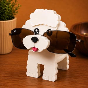 May include: A white dog-shaped sunglasses holder with black eyes, a black nose, and a pink tongue. The holder is wearing a pair of Ray-Ban sunglasses with brown lenses. The holder is sitting on a wooden surface.