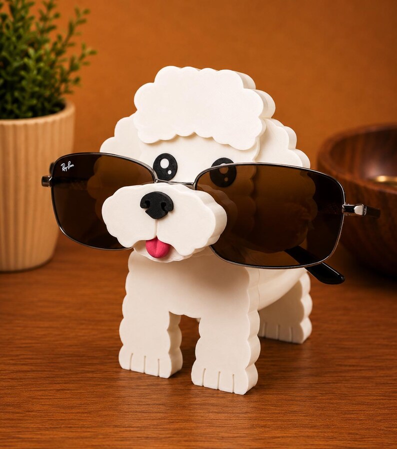 May include: A white dog-shaped sunglasses holder with black eyes, a black nose, and a pink tongue. The holder is wearing a pair of Ray-Ban sunglasses with brown lenses. The holder is sitting on a wooden surface.