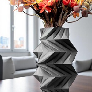 May include: A geometric, gray vase with a zig-zag design holds a bouquet of colorful artificial flowers. The flowers include orange, yellow, and pink hues. The vase sits on a dark wooden table, with a window and a couch in the background.