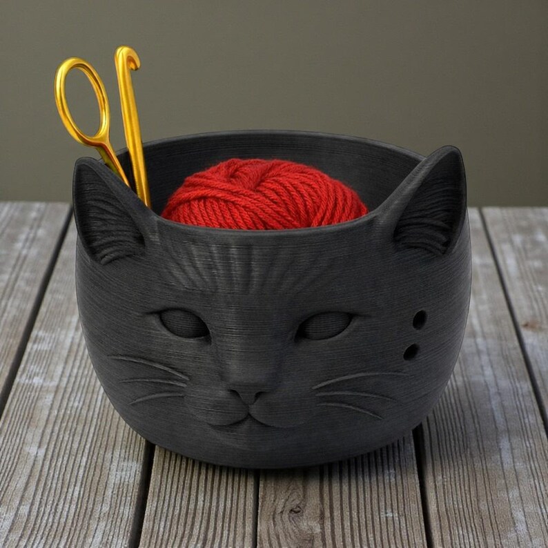 May include: A black cat-shaped yarn bowl containing a red ball of yarn and gold scissors. The bowl has a textured finish and is placed on a wooden surface. The cat's face is detailed with eyes, nose, and whiskers.