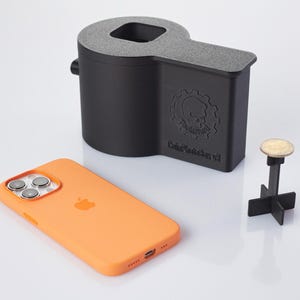 Op de afbeelding: Een zwarte 3D-geprinte telefoonstandaard met een schedel- en tandwielontwerp. De standaard is gelabeld "Data Destroyer V3". Een zwart-witte telefoonhoes ligt op een wit oppervlak naast de standaard. Een kleine zwart-witte standaard met een muntstuk erop staat ook op het oppervlak.