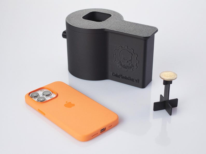 Op de afbeelding: Een zwarte 3D-geprinte telefoonstandaard met een schedel- en tandwielontwerp. De standaard is gelabeld "Data Destroyer V3". Een zwart-witte telefoonhoes ligt op een wit oppervlak naast de standaard. Een kleine zwart-witte standaard met een muntstuk erop staat ook op het oppervlak.