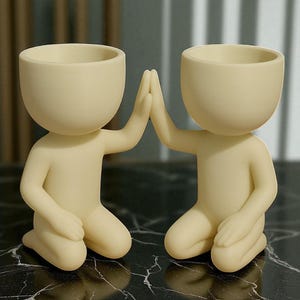 May include: Two cream-colored human-shaped planters. The planters are kneeling with their hands touching in a high-five gesture. The planters have a rounded head and a small opening at the top for plants. They are sitting on a black and white marble surface.