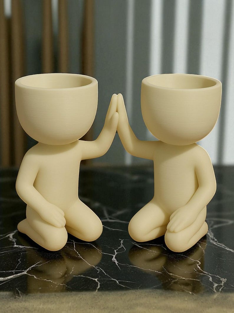 May include: Two cream-colored human-shaped planters. The planters are kneeling with their hands touching in a high-five gesture. The planters have a rounded head and a small opening at the top for plants. They are sitting on a black and white marble surface.