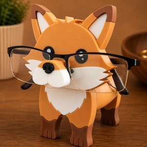 May include: A whimsical orange fox-shaped glasses holder with black-framed glasses. The fox has white and brown accents, a black nose, and large eyes. The glasses are held in place by the fox's nose and mouth, creating a fun and functional accessory.