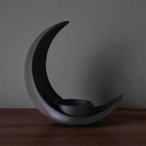 May include: A black ceramic crescent moon candle holder. The holder features a curved design with a circular space to hold a candle. The candle holder is placed on a dark wooden surface.