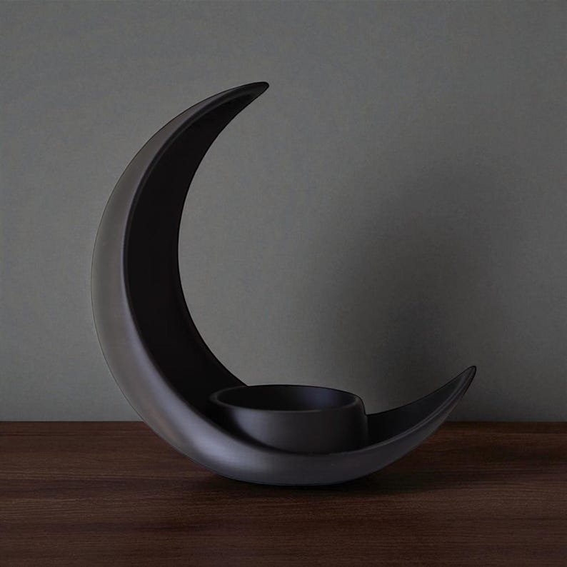 May include: A black ceramic crescent moon candle holder. The holder features a curved design with a circular space to hold a candle. The candle holder is placed on a dark wooden surface.
