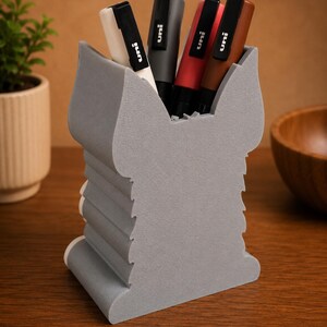May include: A gray pen holder with a unique, layered design, holding several pens. The pens are black, gray, red, and brown, with the brand "uni" visible on each. The holder sits on a wooden surface.