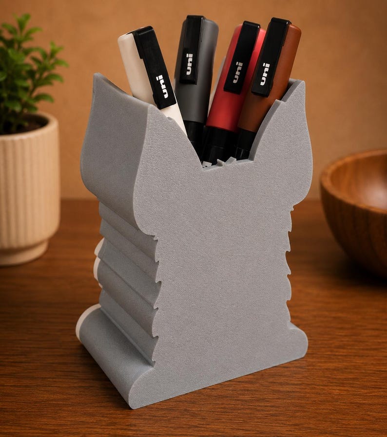 May include: A gray pen holder with a unique, layered design, holding several pens. The pens are black, gray, red, and brown, with the brand "uni" visible on each. The holder sits on a wooden surface.