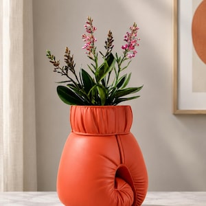 Könnte beinhalten: Eine leuchtend orangefarbene Vase in Form eines Boxhandschuhs mit gerafftem Bündchen am oberen Rand. Die Vase enthält eine kleine grüne Pflanze mit dünnen Stielen und zarten rosa-weißen Blüten, die einen Kontrast zur glatten Oberfläche bildet.