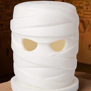 K&ouml;nnte beinhalten: Ein wei&szlig;er, zylindrischer dekorativer Kerzenhalter in Form einer Mumie. Das Gesicht der Mumie hat zwei gro&szlig;e, ovale Augenl&ouml;cher, die mit einem warmen, gelben Licht leuchten. Das Design zeigt Wicklungen um Kopf und K&ouml;rper, was ein gruseliges Halloween-Thema erzeugt.