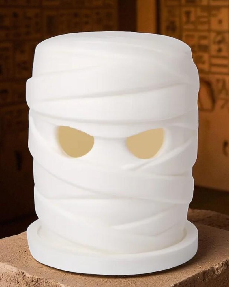 K&ouml;nnte beinhalten: Ein wei&szlig;er, zylindrischer dekorativer Kerzenhalter in Form einer Mumie. Das Gesicht der Mumie hat zwei gro&szlig;e, ovale Augenl&ouml;cher, die mit einem warmen, gelben Licht leuchten. Das Design zeigt Wicklungen um Kopf und K&ouml;rper, was ein gruseliges Halloween-Thema erzeugt.