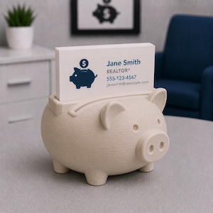 May include: A beige piggy bank business card holder with a slot for cards. The holder has a pig shape with ears, a snout, and four legs. Business cards with the name "Jane Smith, REALTOR" are in the holder.