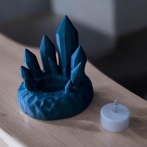 May include: A teal crystal-shaped candle holder with a central well and surrounding pointed crystal formations. A small, white LED tea light sits beside the holder. The holder is made of a textured material and sits on a wooden surface.