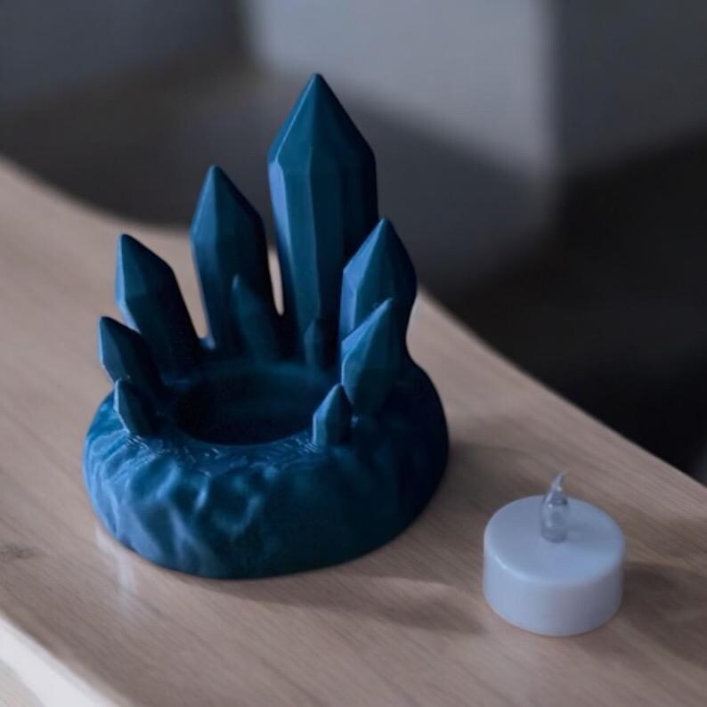 May include: A teal crystal-shaped candle holder with a central well and surrounding pointed crystal formations. A small, white LED tea light sits beside the holder. The holder is made of a textured material and sits on a wooden surface.