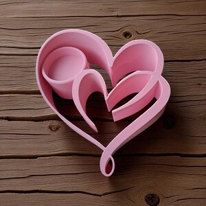 May include: A pink heart-shaped candle holder with a nested design. The holder features a central circular cup and a larger heart outline, all in a soft pink hue. The design is set against a wooden background.
