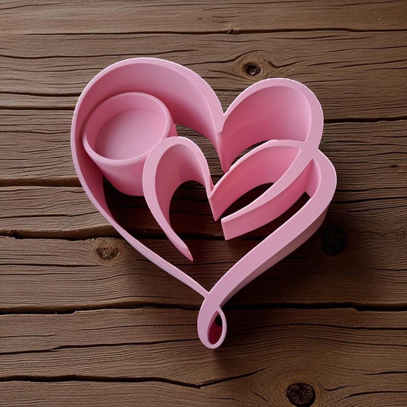 May include: A pink heart-shaped candle holder with a nested design. The holder features a central circular cup and a larger heart outline, all in a soft pink hue. The design is set against a wooden background.