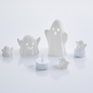 May include: Set of five white ceramic ghost candle holders with different facial expressions. The ghosts are various sizes and are lit with tea lights.
