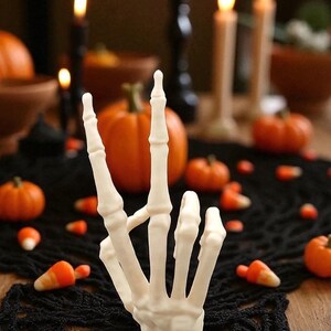 May include: A white, skeletal hand sculpture giving a peace sign. The Halloween-themed decoration is surrounded by miniature pumpkins, candy corn, and lit candles on a black lace tablecloth. The background is blurred.