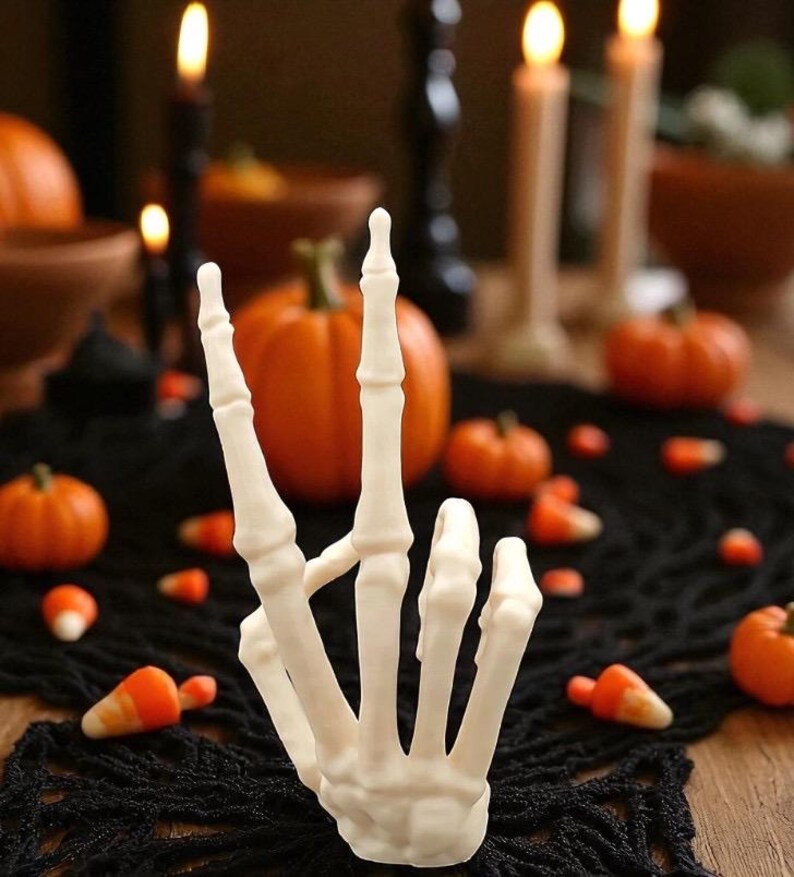 May include: A white, skeletal hand sculpture giving a peace sign. The Halloween-themed decoration is surrounded by miniature pumpkins, candy corn, and lit candles on a black lace tablecloth. The background is blurred.