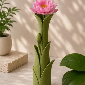 以下が含まれることがあります： 黄色い中心部を持つピンク色の蓮の花が入った、緑色の葉の形をした花瓶。花瓶の側面にはつぼみがあります。背景はニュートラルカラーで、背景には他の植物があります。