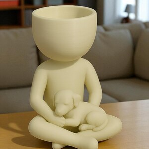 May include: A cream-colored decorative planter shaped like a person seated with legs crossed, cradling a sleeping puppy. The planter has a large, open head for planting. The figurine is made of a smooth material and sits on a wooden surface.