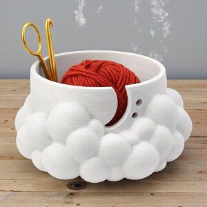 K&ouml;nnte beinhalten: Eine wei&szlig;e Garnschale mit einem wolkenartigen Design, die einen roten Wollkn&auml;uel und eine goldene H&auml;kelnadel und Schere enth&auml;lt. Die Schale hat eine kreisf&ouml;rmige &Ouml;ffnung f&uuml;r das Garn und zwei kleine L&ouml;cher. Die Schale steht auf einer Holzoberfl&auml;che.