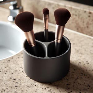 Peut inclure: Un porte-pinceaux gris foncé à trois compartiments contenant trois pinceaux de maquillage. Les pinceaux ont des poils brun foncé et des manches couleur or rose. Le support est posé sur un comptoir moucheté.