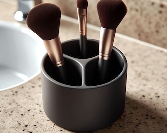 May include: A dark gray makeup brush holder with three compartments, holding three makeup brushes. The brushes have dark brown bristles and rose gold-colored handles. The holder sits on a speckled countertop.