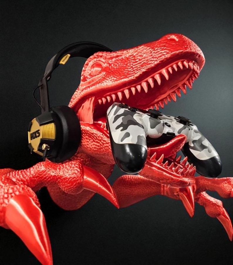 May include: A red dinosaur sculpture holding a camouflage-patterned game controller. A black and yellow headset rests on the dinosaur's head. The headset has the text "HXS" on the side.