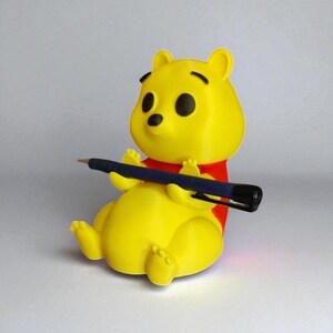 May include: A yellow figurine of a bear holding a blue pen. The bear has black eyes and a black nose, and is wearing a red shirt. The figurine is sitting down with its legs extended.