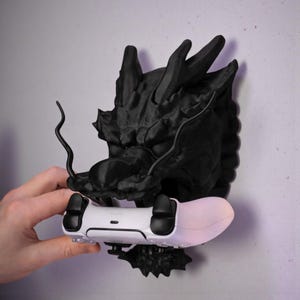 May include: A black dragon head wall mount holding a white and black game controller. The dragon head has detailed features and is mounted on a light-colored wall. The controller is held in place by the dragon's mouth.