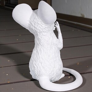 May include: A white sculpture of a mouse with oversized ears and a long, curved tail. The mouse is depicted from the back, with its hand raised near its head. The sculpture has a textured surface and is set against a neutral background.