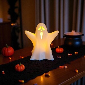 May include: A glowing white ghost-shaped Halloween decoration with outstretched arms. The ghost has yellow eyes and a mouth, and is lit from within. Small orange pumpkins and candy corn are scattered on a black runner.