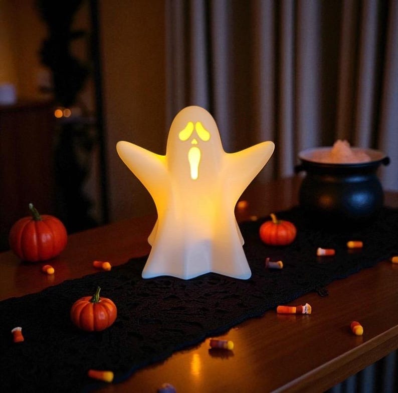 May include: A glowing white ghost-shaped Halloween decoration with outstretched arms. The ghost has yellow eyes and a mouth, and is lit from within. Small orange pumpkins and candy corn are scattered on a black runner.