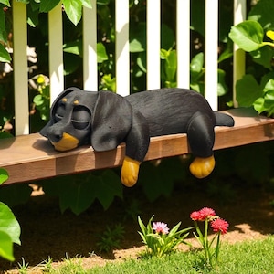 Pu&ograve; includere: Una statua di cane bassotto nero e marrone chiaro &egrave; appoggiata su una panchina di legno. Il cane ha gli occhi chiusi e le zampe gialle. La statua si trova in un giardino con fiori e una staccionata bianca sullo sfondo.