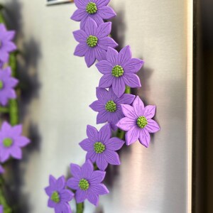 May include: A garland of purple flower magnets with green centers and green stems. The flowers are attached to a metallic surface, creating a decorative floral arrangement. The magnets are a vibrant addition to any kitchen or office.