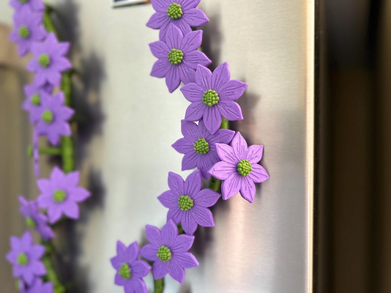May include: A garland of purple flower magnets with green centers and green stems. The flowers are attached to a metallic surface, creating a decorative floral arrangement. The magnets are a vibrant addition to any kitchen or office.