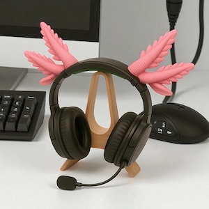 Puede incluir: Auriculares negros de diadema con micr&oacute;fono, adornados con detalles rosas inspirados en ajolotes. Los auriculares descansan sobre un soporte de madera. Un teclado y un rat&oacute;n de ordenador se ven al fondo. Para juegos o audio.