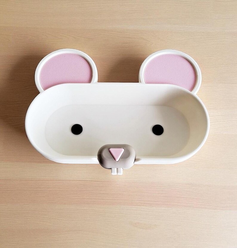 May include: A white, oval-shaped soap dish with a mouse face design. It features two pink ears, black eyes, and a gray nose. The dish is made of plastic and sits on a light brown wooden surface.
