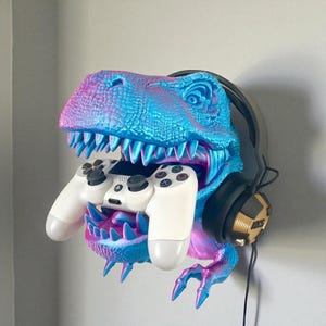 Könnte beinhalten: Eine farbenfrohe, an der Wand befestigte Dinosaurierkopf-Skulptur in Blau-, Rosa- und Lilatönen, die einen weißen Gamecontroller hält. Schwarze Kopfhörer mit goldenen Akzenten sind an der Seite positioniert. Die Skulptur dient zur Aufbewahrung von Gaming-Zubehör.