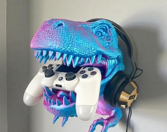 May include: A vibrant, wall-mounted dinosaur head sculpture in shades of blue, pink, and purple, holding a white game controller. Black headphones with gold accents are positioned on the side. The sculpture is designed to hold gaming accessories.
