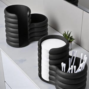 Pode incluir: Dois organizadores de casa de banho pretos e modernos com um design &uacute;nico e canelado. Um cont&eacute;m discos de algod&atilde;o, o outro cotonetes. Os organizadores est&atilde;o sobre uma bancada branca, com uma pequena planta no fundo.